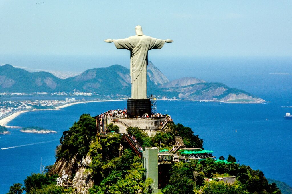 Cristo Redentor de Braços abetos, foto vista do alto. Pessoas visitando a estátua. Dia de sol, mar azul.