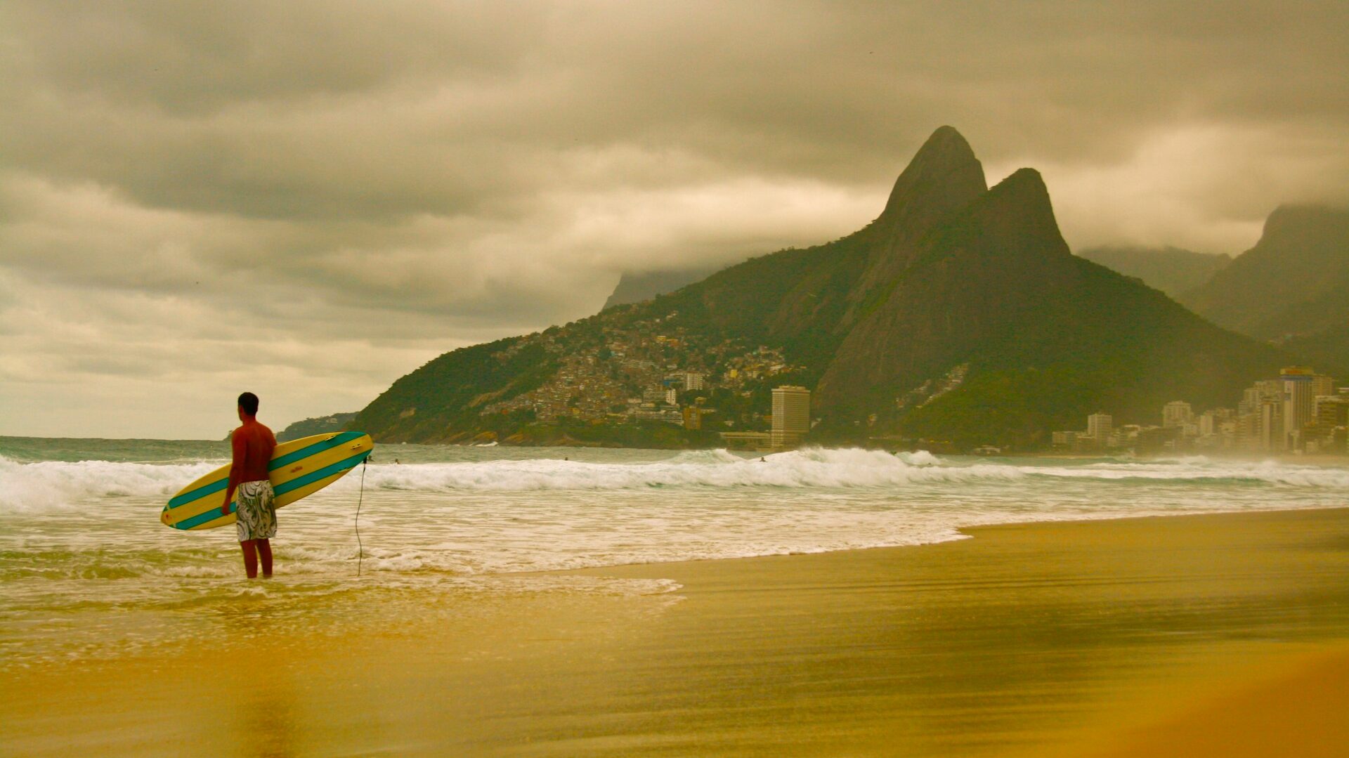 Foto praia de Ipanema, fim de tarde, céu nublado. Surfista segurando prancha olhando as ondas.