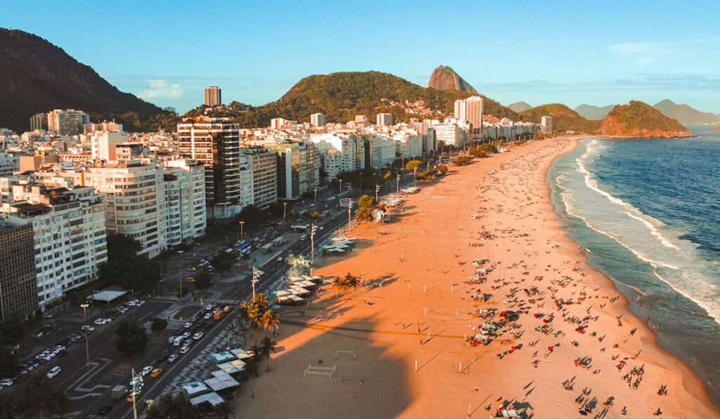 Foto em dia de sol da areia e mar da praia de Copacabana, Rio de Janeiro; Pessoas na areia.